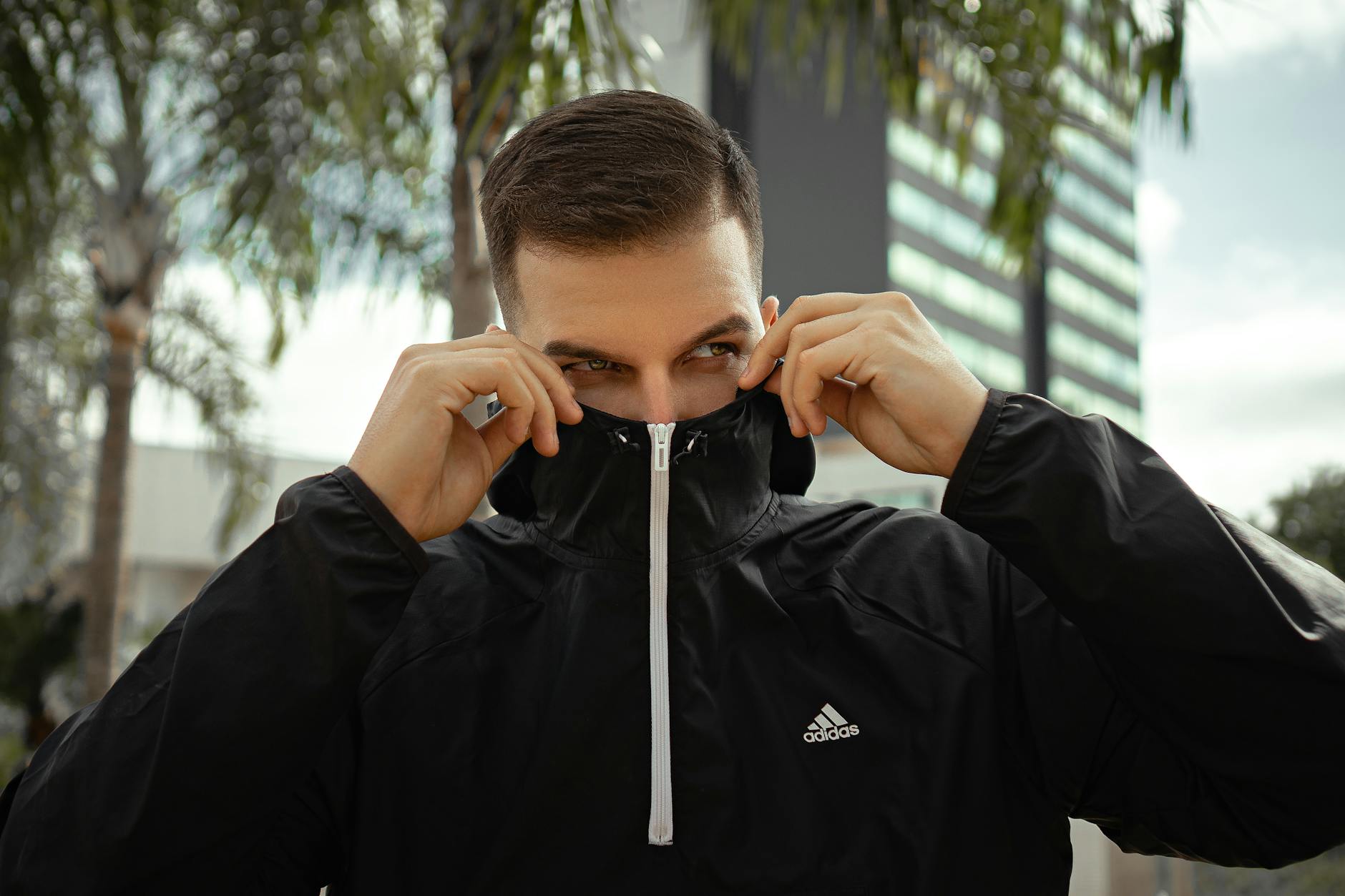 Young man adjusts hood of black Adidas jacket outdoors, contemporary urban setting.