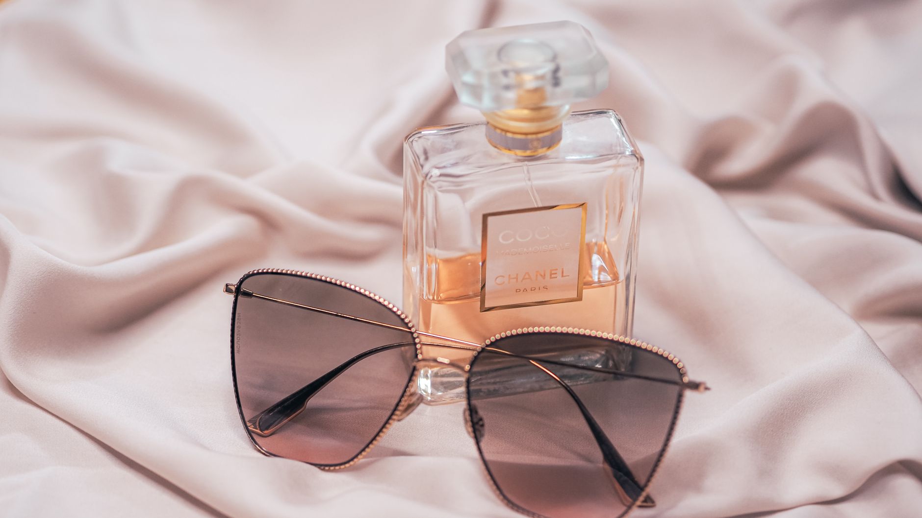Stylish Coco Chanel perfume and sunglasses arrangement on silk fabric.